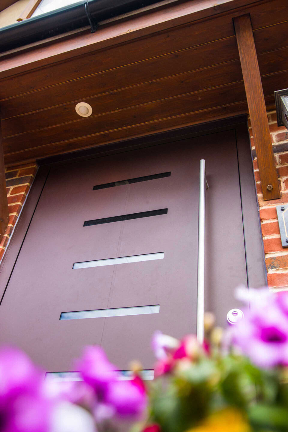Close-up view of a brown aluminium front door with three slim horizontal glass inserts and a vertical pull handle, set in a brick exterior.