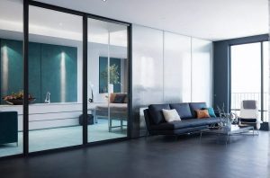 Modern living room featuring a dark hardwood floor and a large glass and black steel-look aluminium internal screen separating the space. A sleek dark grey sofa adorned with various decorative pillows sits opposite a minimalist glass coffee table with an array of glassware and a serving tray. Beyond the screen, a kitchen area with teal walls and white countertops is partially visible. Natural light filters through the large windows and balcony door, highlighting the tasteful mix of contemporary and cosy décor elements.