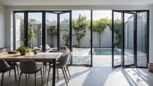 Modern indoor dining area with a wooden table and grey chairs, opening to an outdoor patio with a pool through sleek aluminium bi-fold doors.