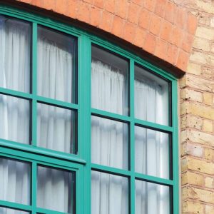 Green-framed arched window set in a brick wall with white curtains behind the glass.