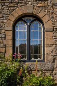 Double arched window in a stone wall with flowers in the foreground, set in a rustic building.