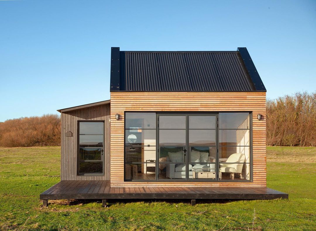 Front view of a timber-clad cabin with large steel look aluminium doors and multi-pane glazing, opening onto a small deck in a grassy field.