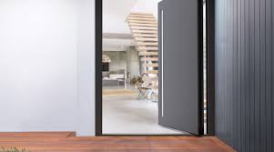 Grey aluminium pivot door with a narrow vertical glazed strip, partially open to reveal a modern interior with wooden staircase.