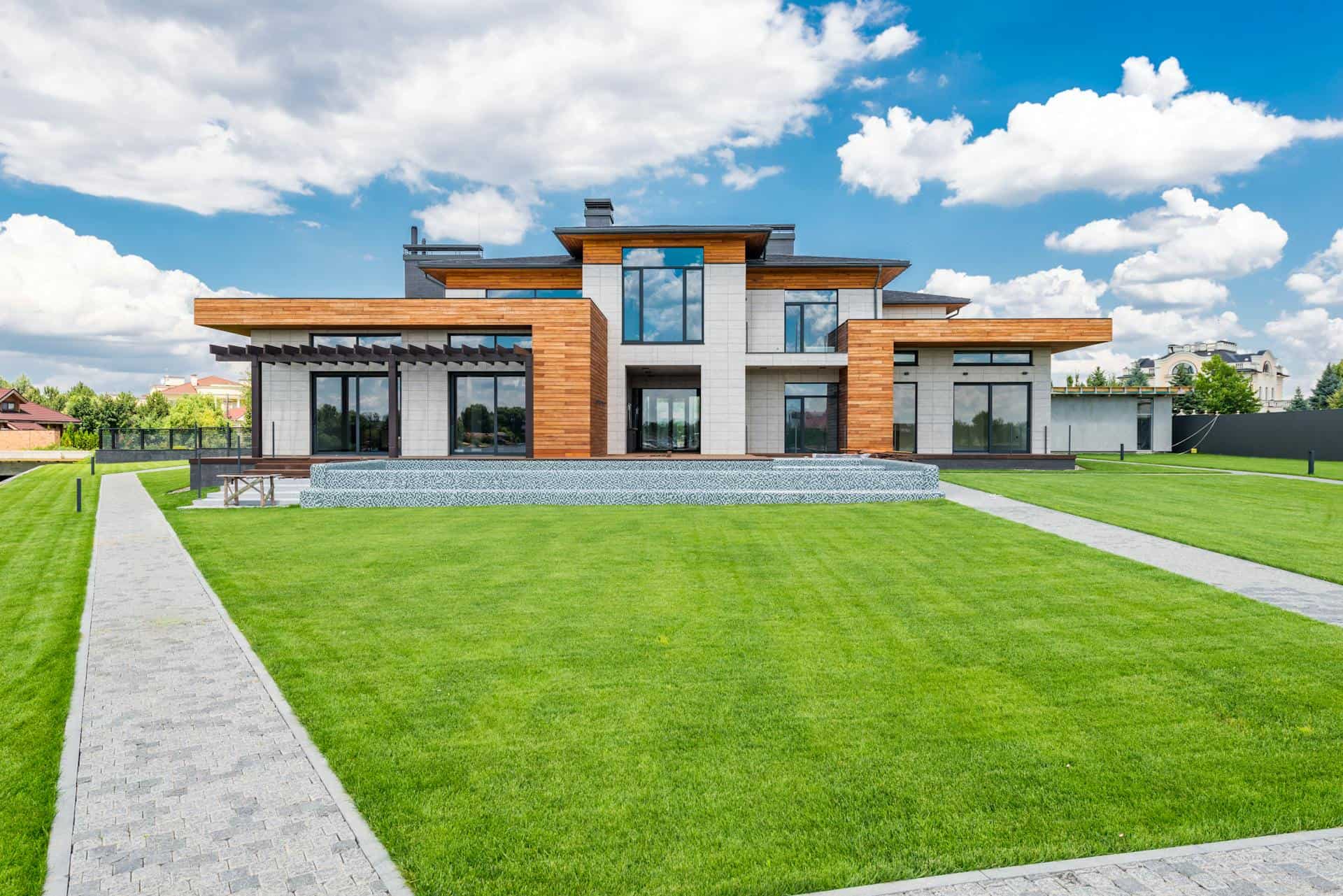 Modern house with large glass windows and wooden accents, set in a lush green garden under a blue sk.